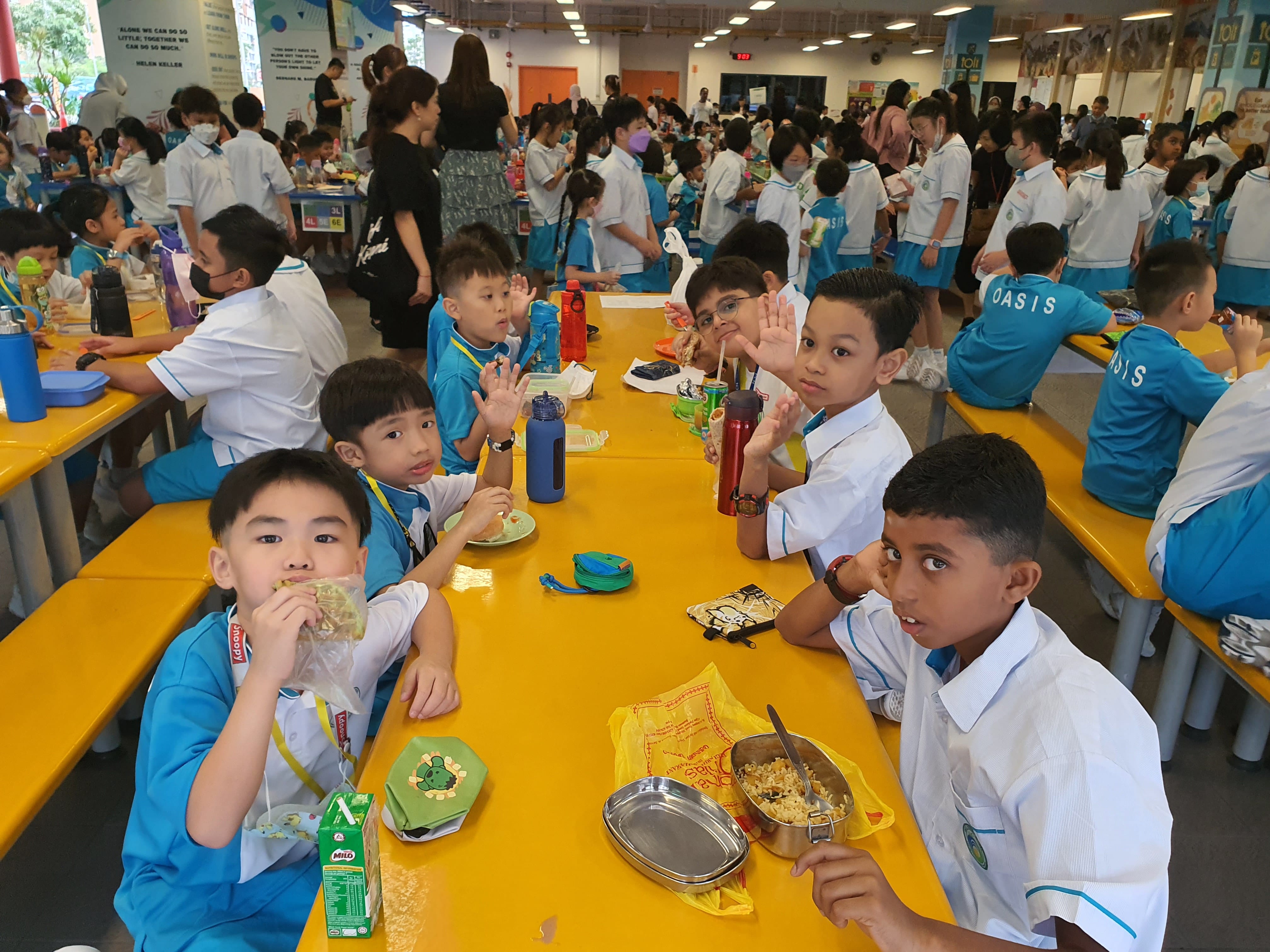 Young students in blue and white uniforms eating lunch at yellow tables in a busy school cafeteria.