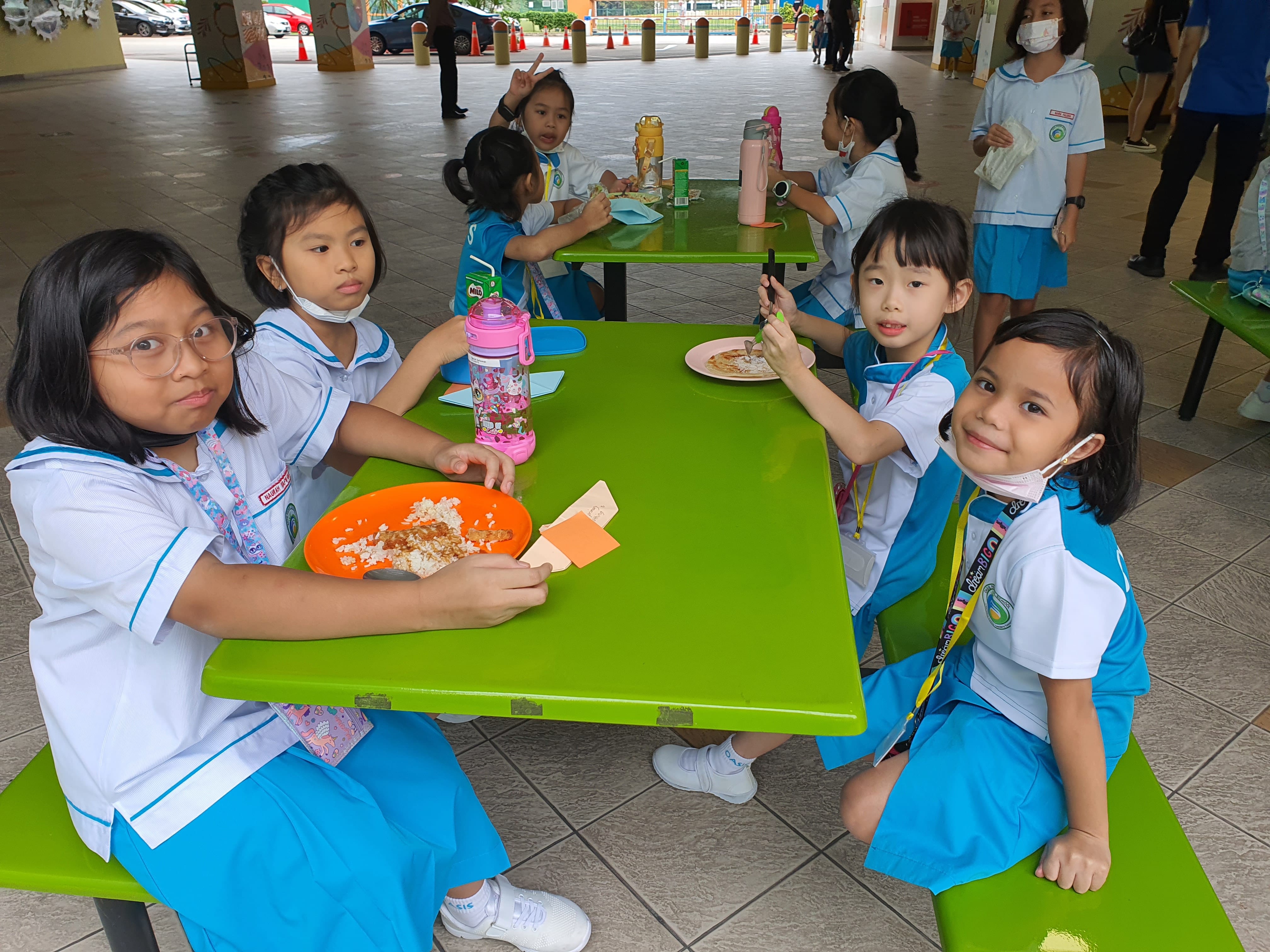 Four young girls in school uniforms sit eating lunch at a bright green table in an outdoor school canteen.