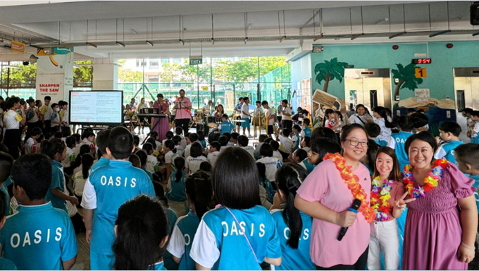 Students in blue Oasis uniforms gather in a school hall while adults wearing flower leis smile near a microphone.