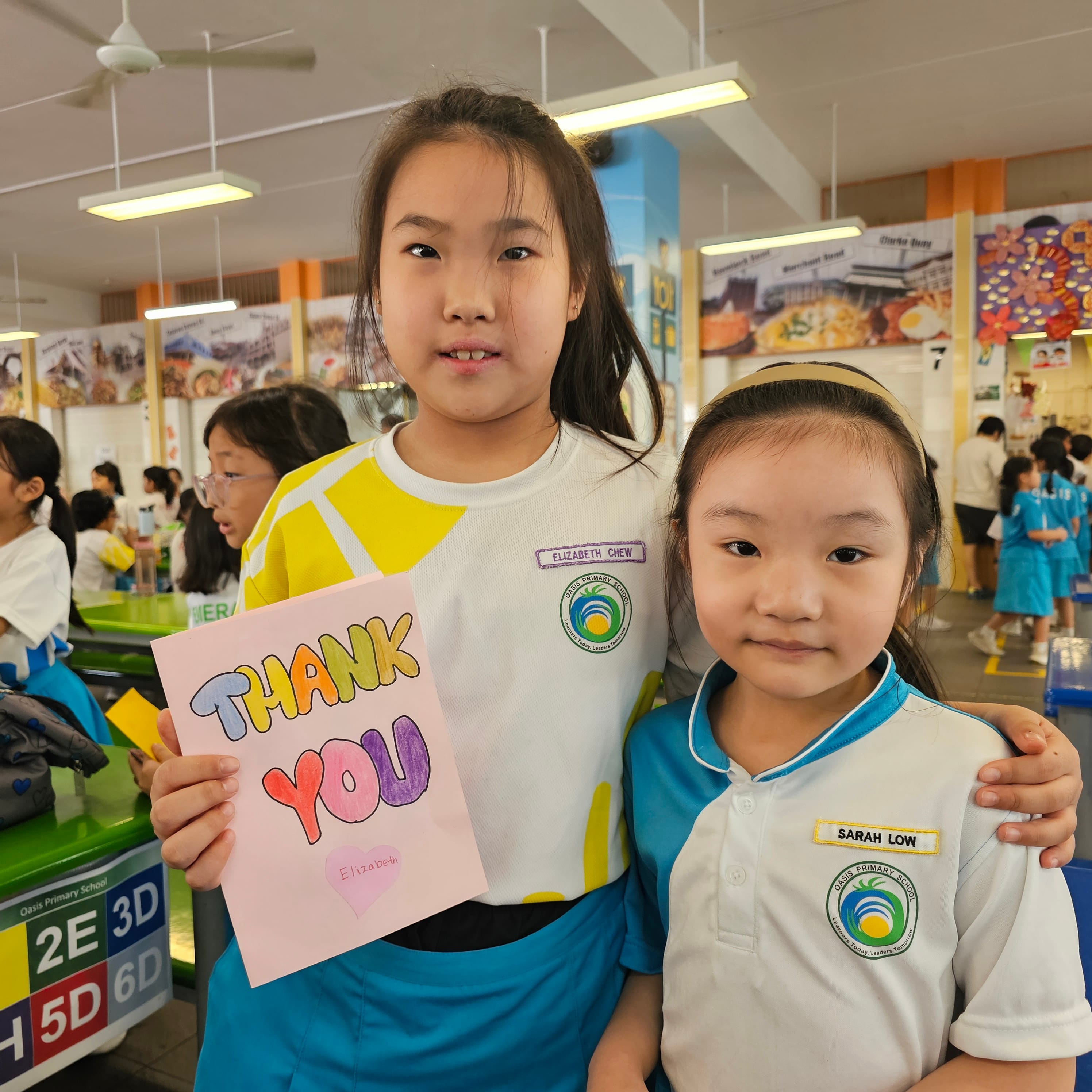 Two girls in Oasis Primary School uniforms pose together, one holding a colorful handmade Thank You card signed Elizabeth.