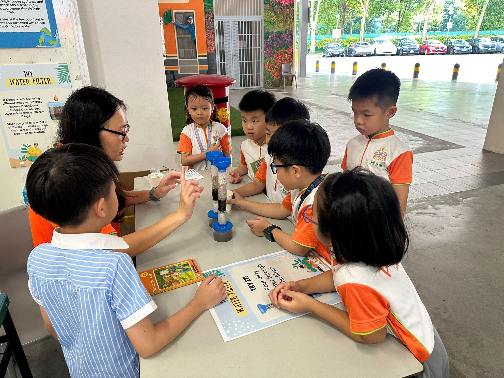 A teacher explains a DIY water filter experiment to young students in orange and white uniforms at a table.