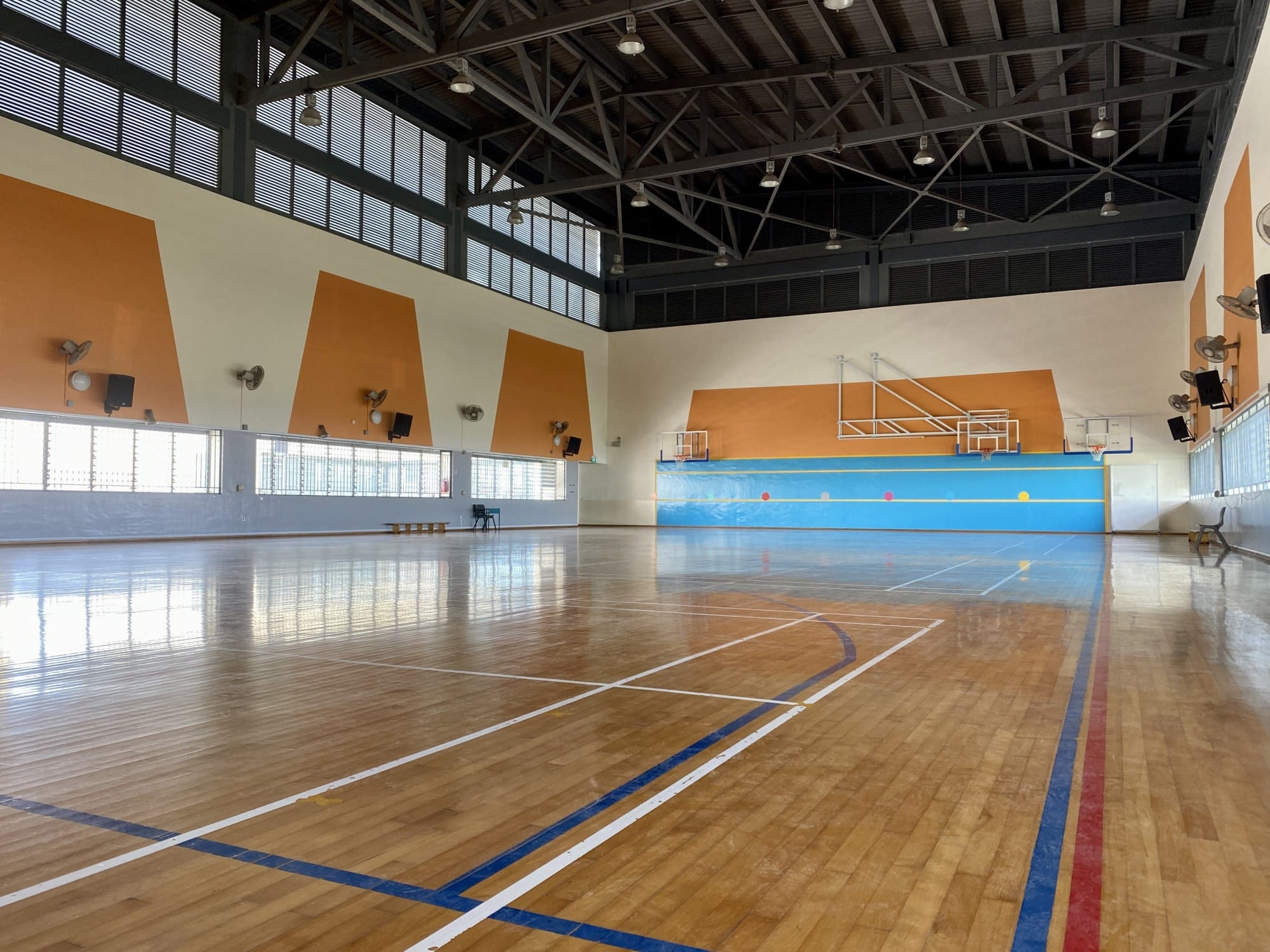 Empty indoor basketball court with wooden floor, orange wall accents, hoops at far end, and steel truss ceiling with hanging lights.