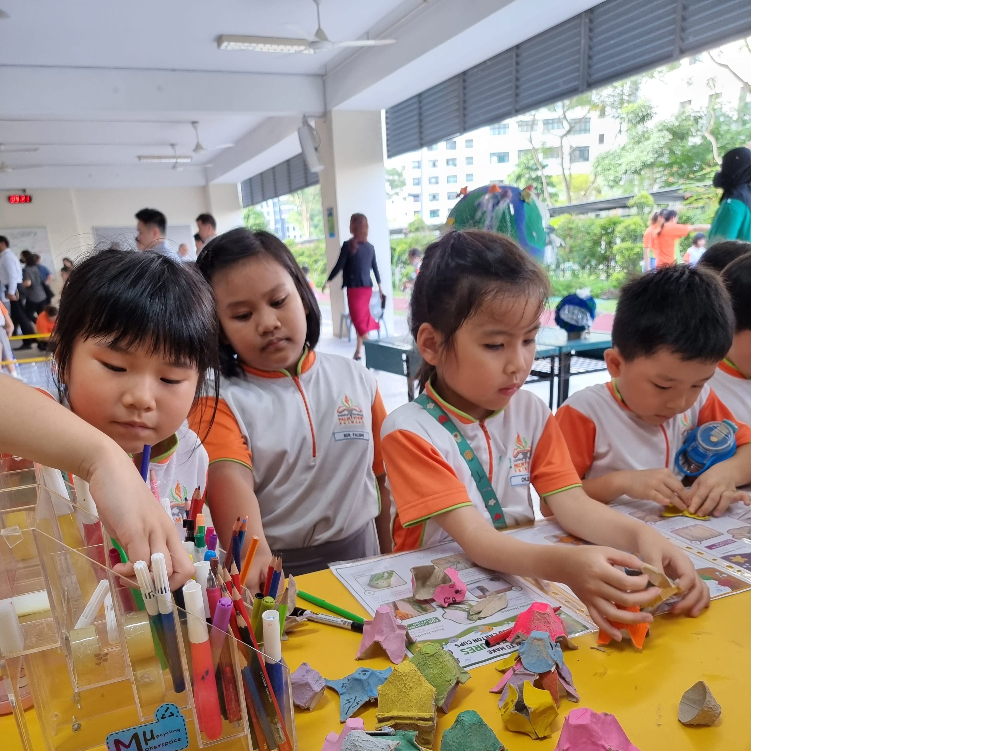 Four young children in school uniforms do a craft activity at a yellow table with egg cartons, markers, and tape.
