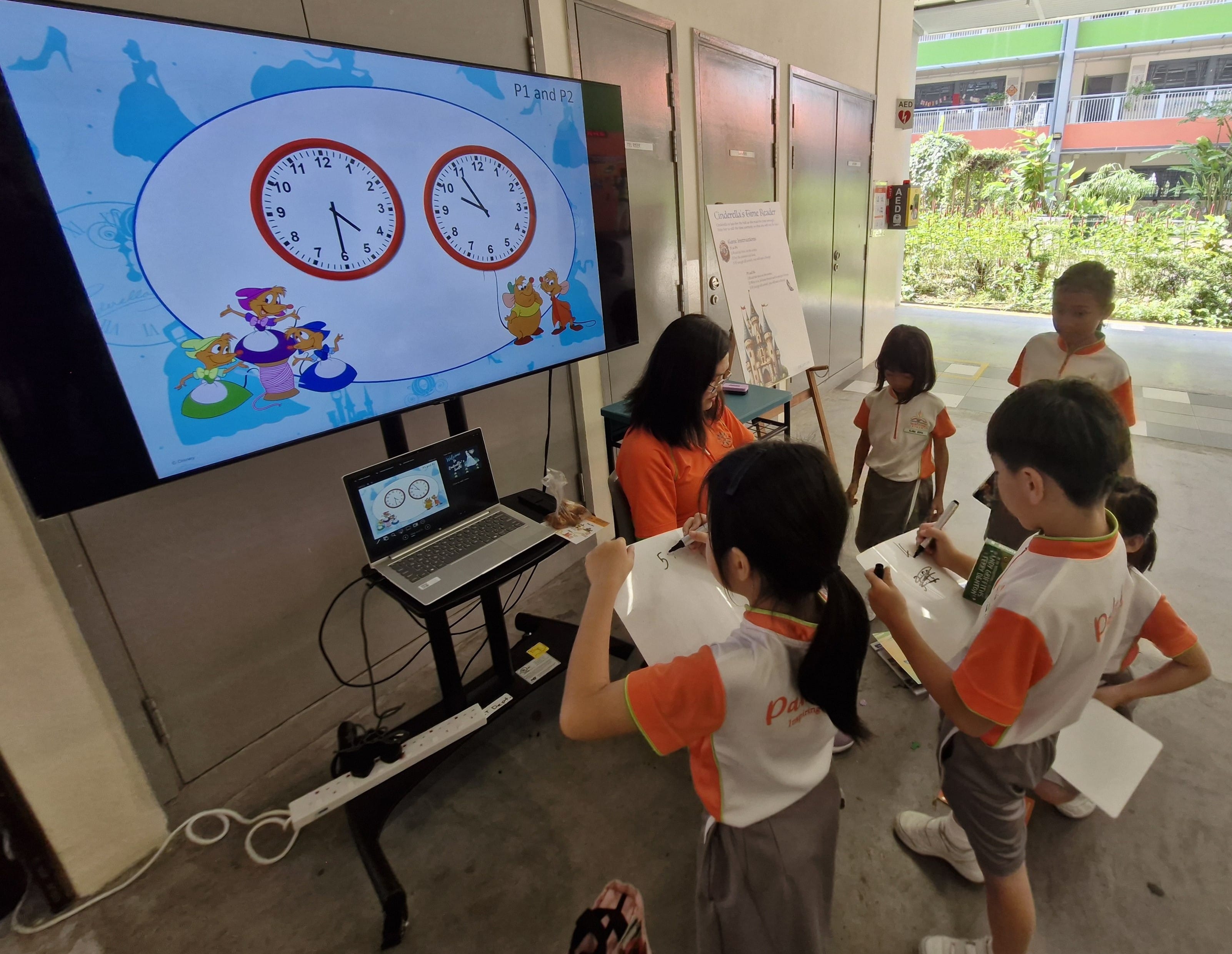 A teacher and young students in orange uniforms do a clock-reading activity using a large screen showing cartoon clocks.