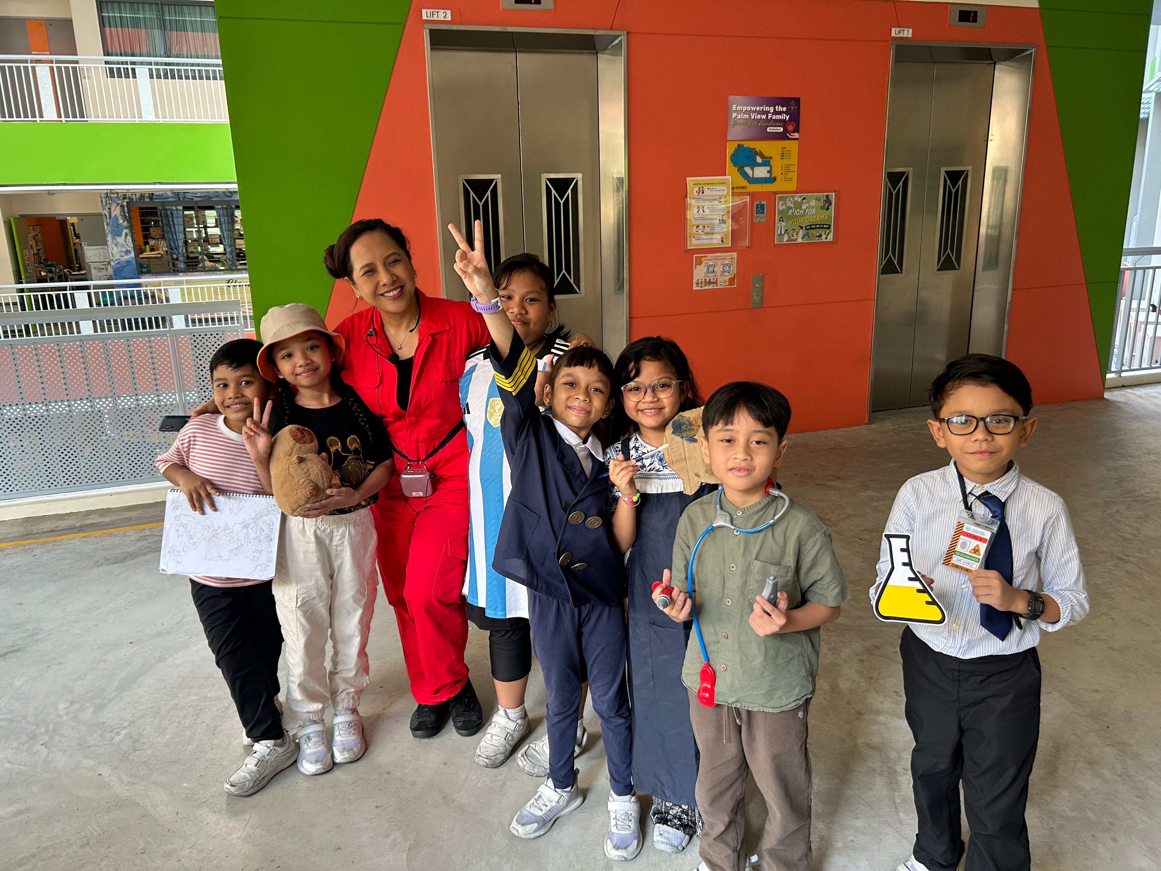 A woman in red and six children in various costumes pose smiling near elevators in a colorful building.