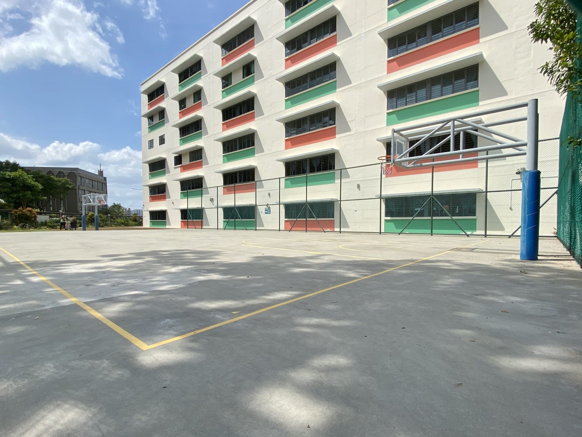 An empty outdoor basketball court with yellow lines beside a multi-story building with red and green accents.