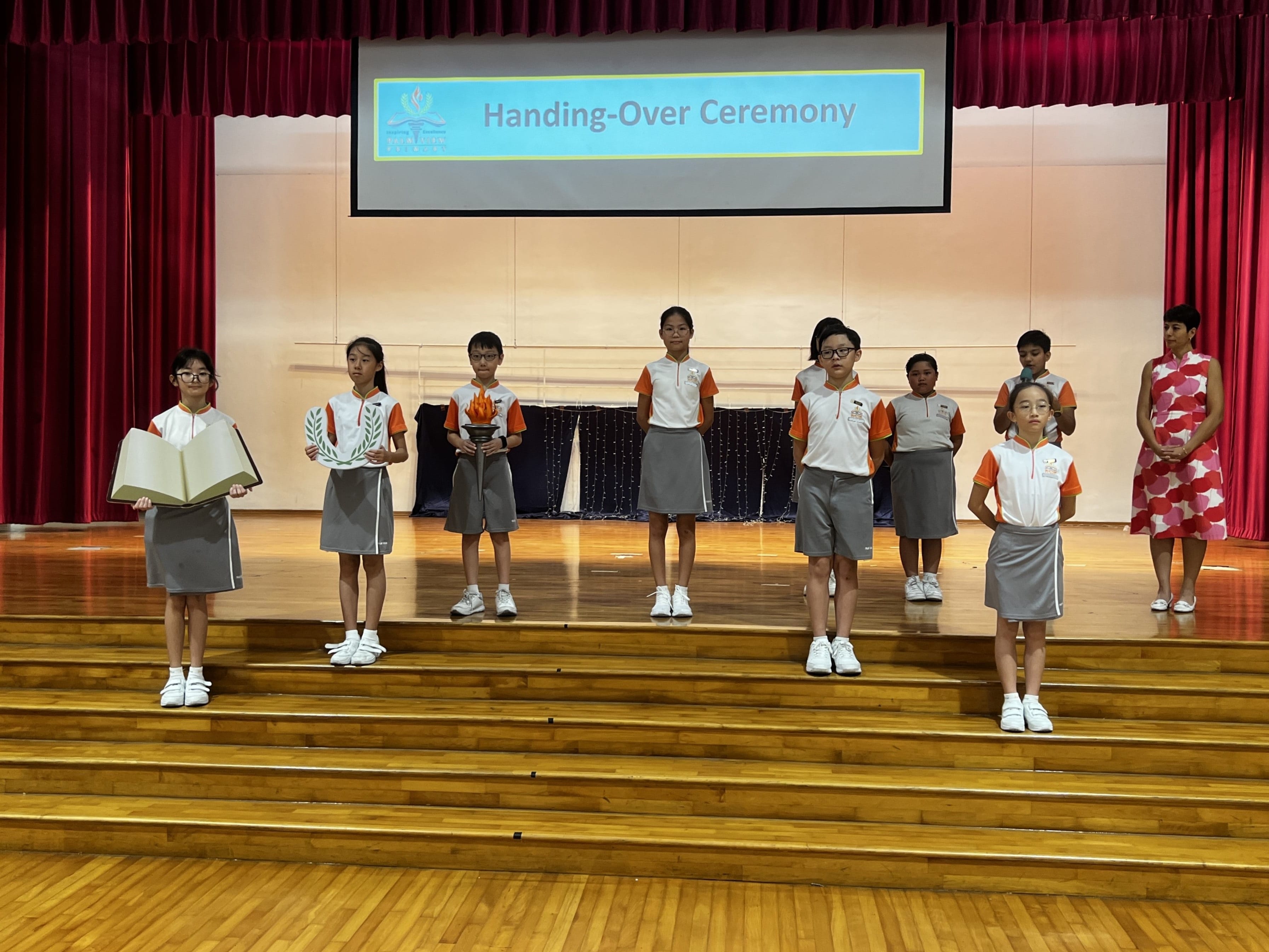 Students in orange and white uniforms stand on a school stage during a Handing-Over Ceremony, holding a torch and book props.