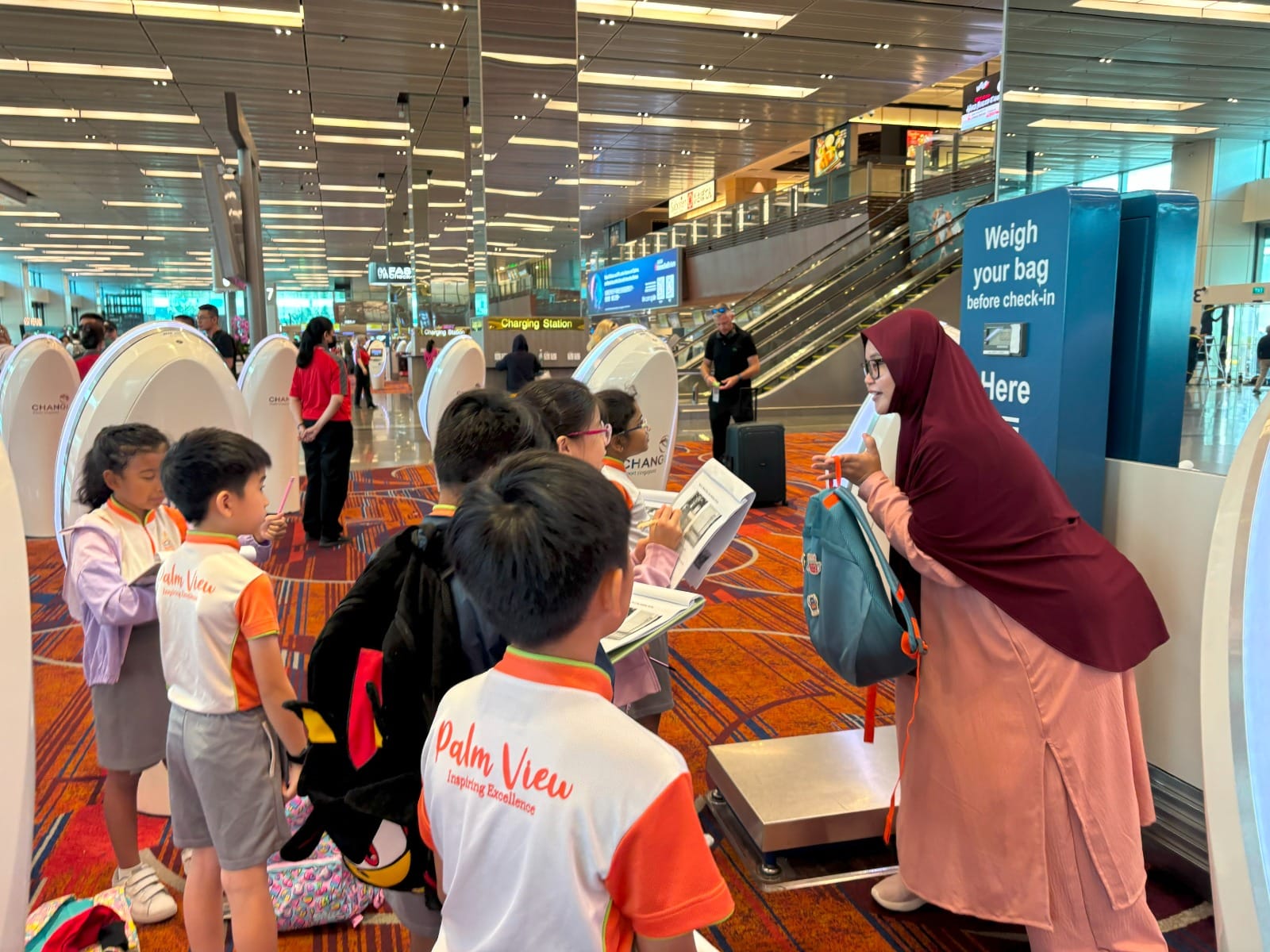 A woman in a hijab guides Palm View school children near a bag weighing station inside Changi Airport.