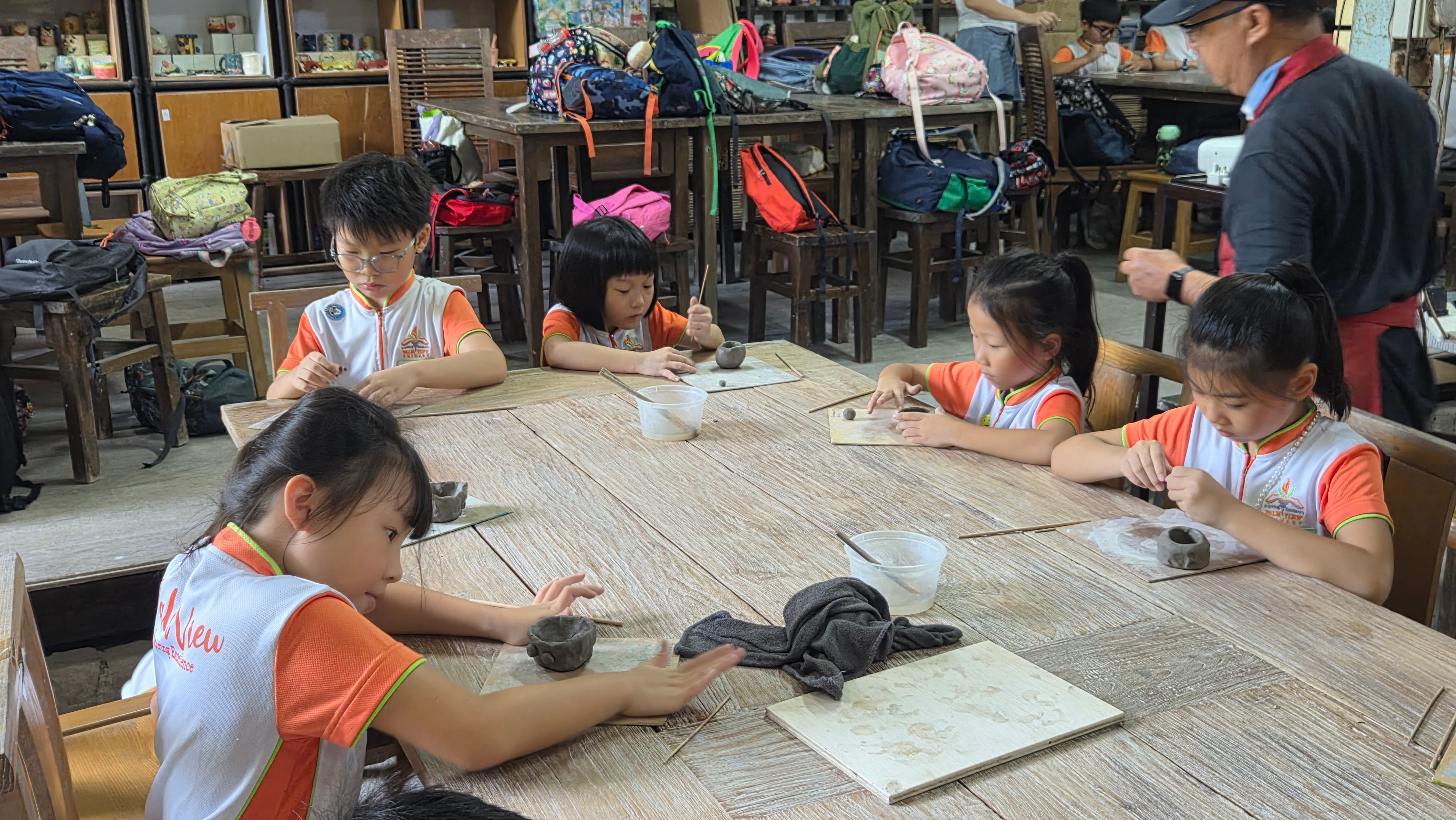 Four young children in matching uniforms shape clay at a wooden table during a pottery class.