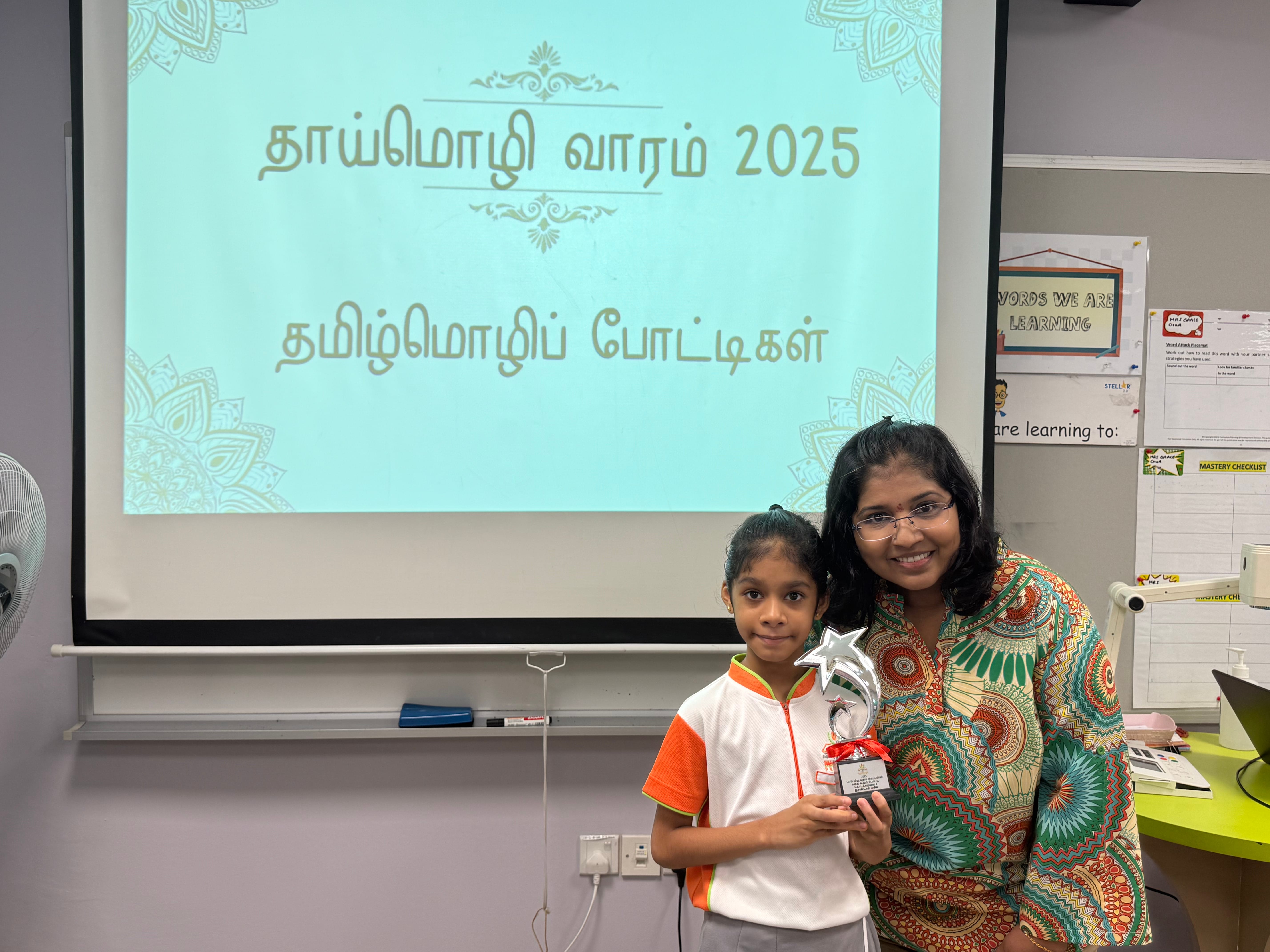 A young girl and a woman hold a star trophy in a classroom before a Tamil language event slide reading 2025.