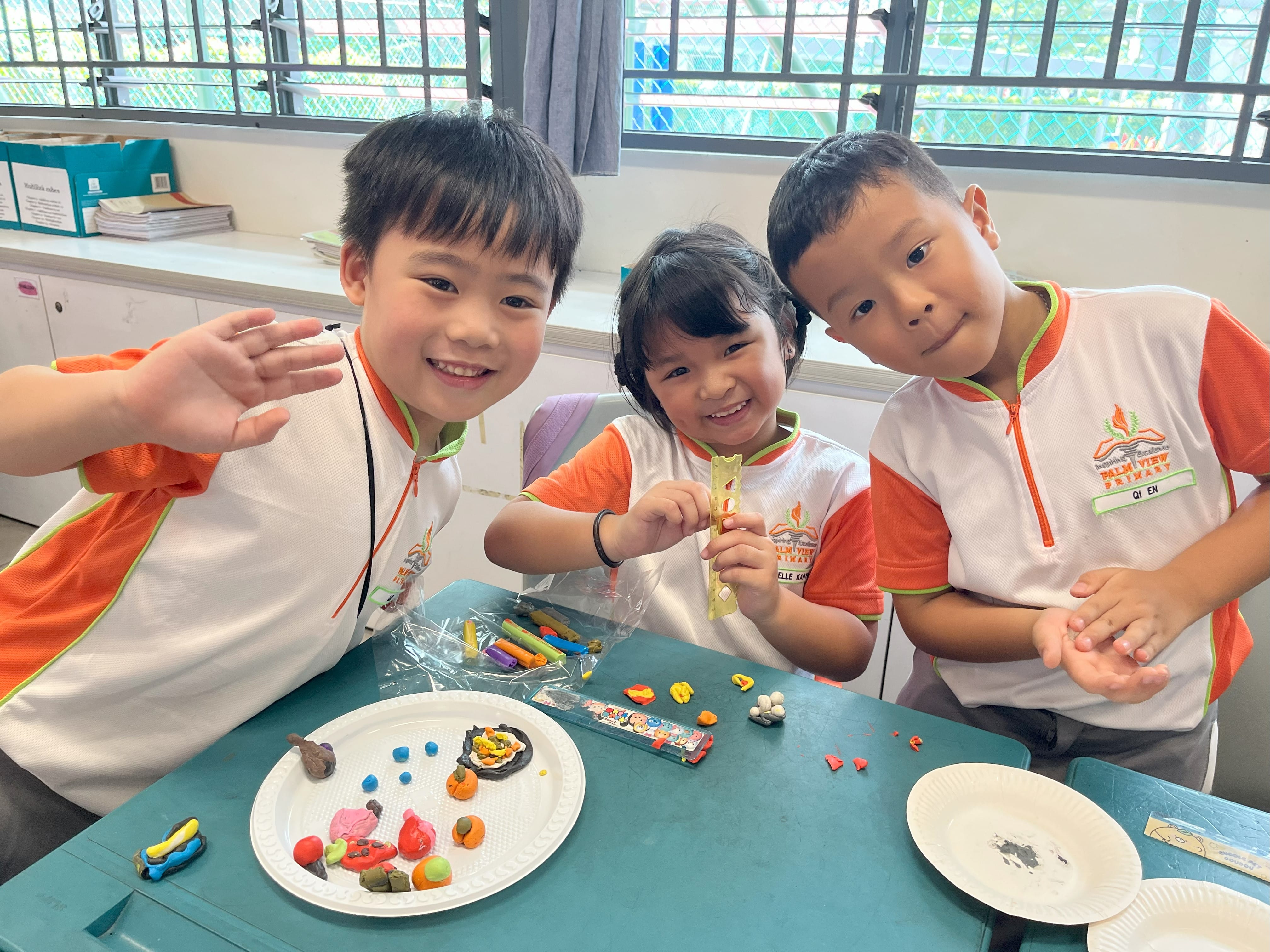 Three young students in matching uniforms smile at a table covered with colorful clay creations on a white plate.
