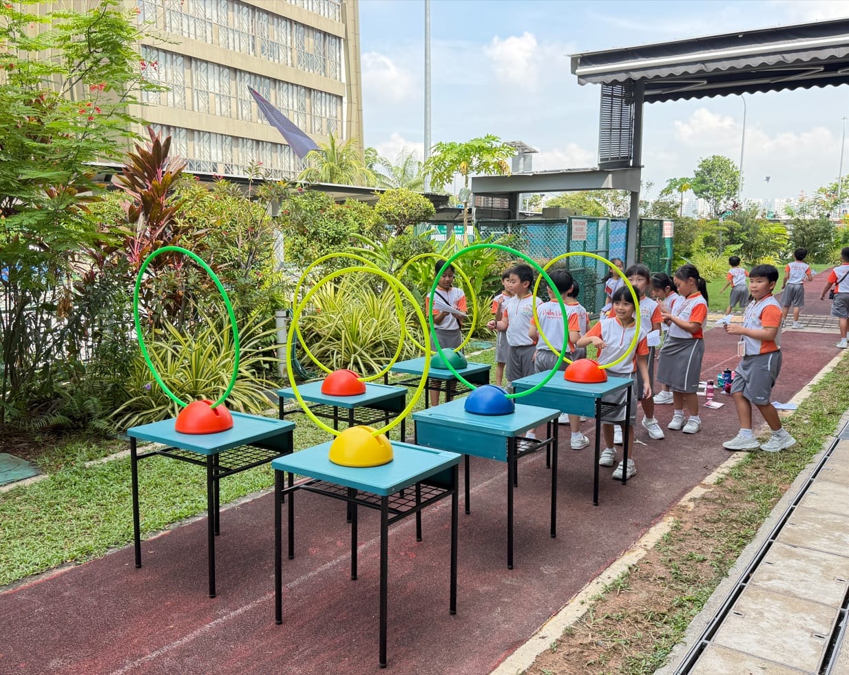 Young students in uniforms stand near tables with colorful dome markers and hula hoops outdoors on a track.