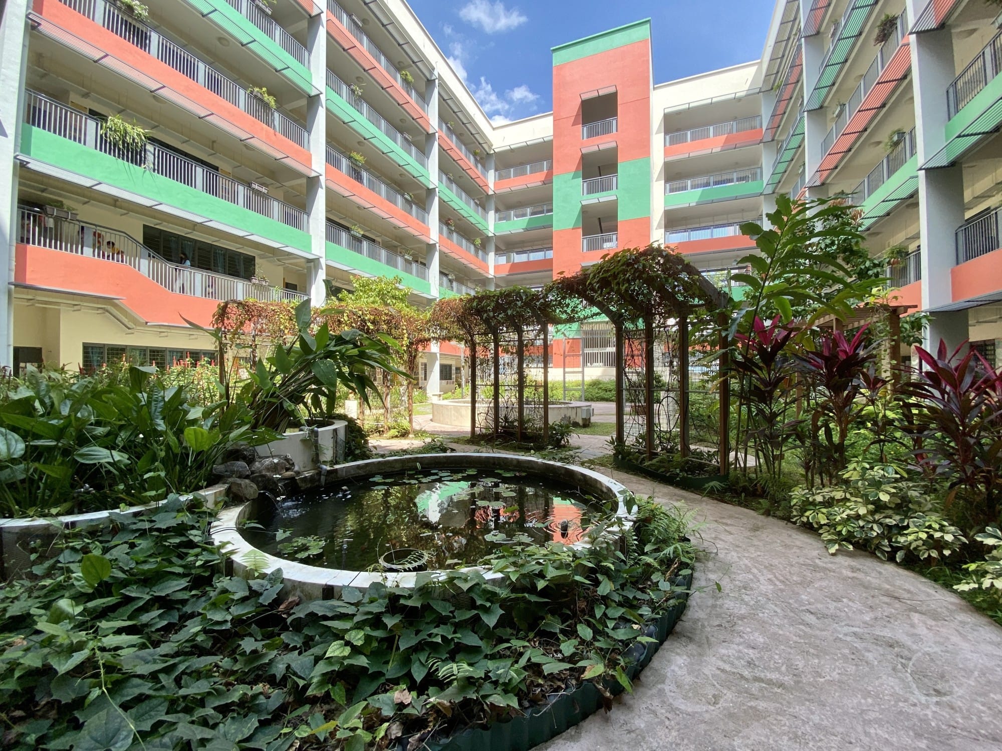Courtyard of a colorful orange and green apartment complex with a round pond, lush plants, and a vine-covered archway path.