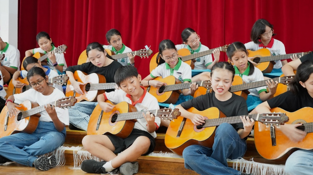 A large group of children in school uniforms sitting on steps, each playing an acoustic guitar before a red curtain.