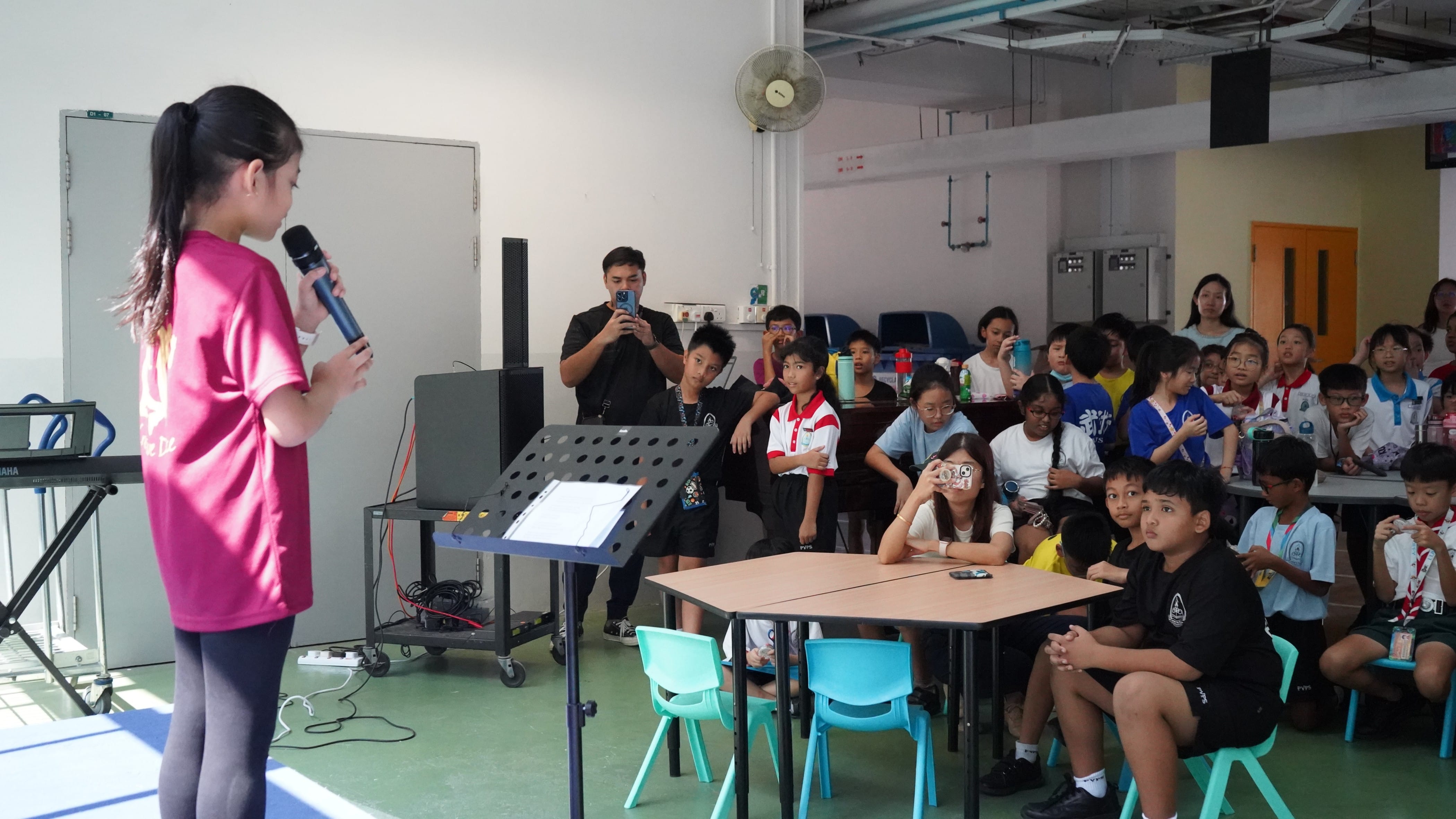 A girl in a pink shirt holds a microphone before a music stand, facing a crowd of seated children in a school hall.