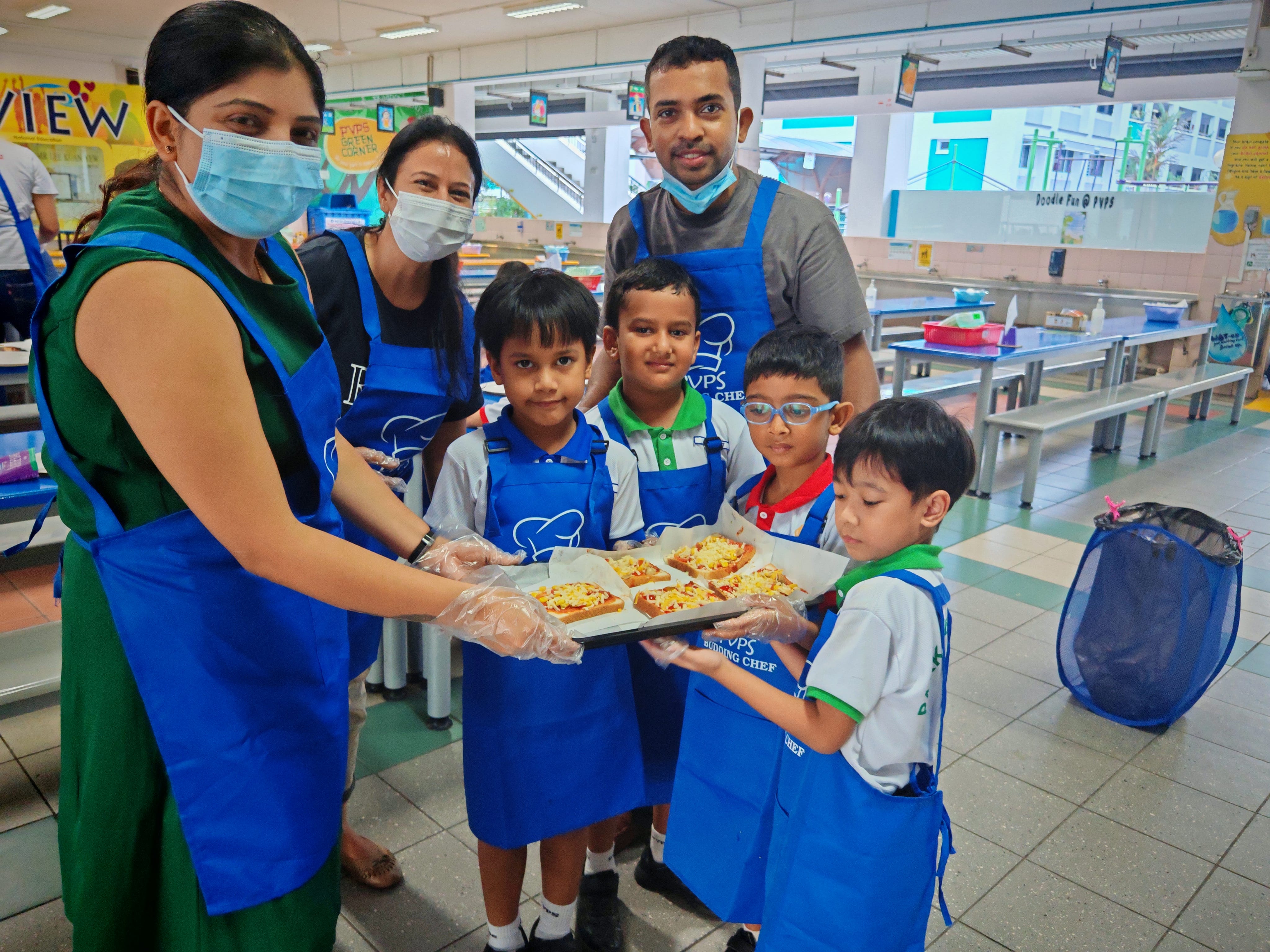 P1 students carrying a tray of mini pizzas with the help of parent volunteers