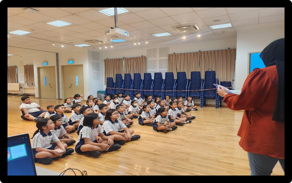 A teacher in red stands before a large group of young students in uniforms sitting on a wooden hall floor.
