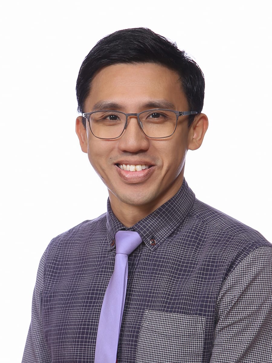 A smiling man with glasses wearing a gray checkered shirt and purple tie against a white background.