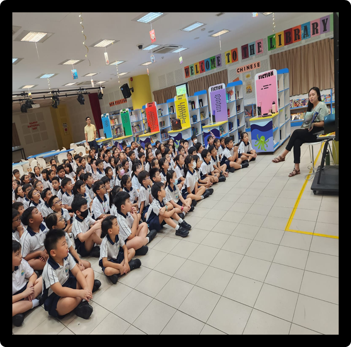 A large group of uniformed students sit on a library floor listening to a teacher seated at the front.
