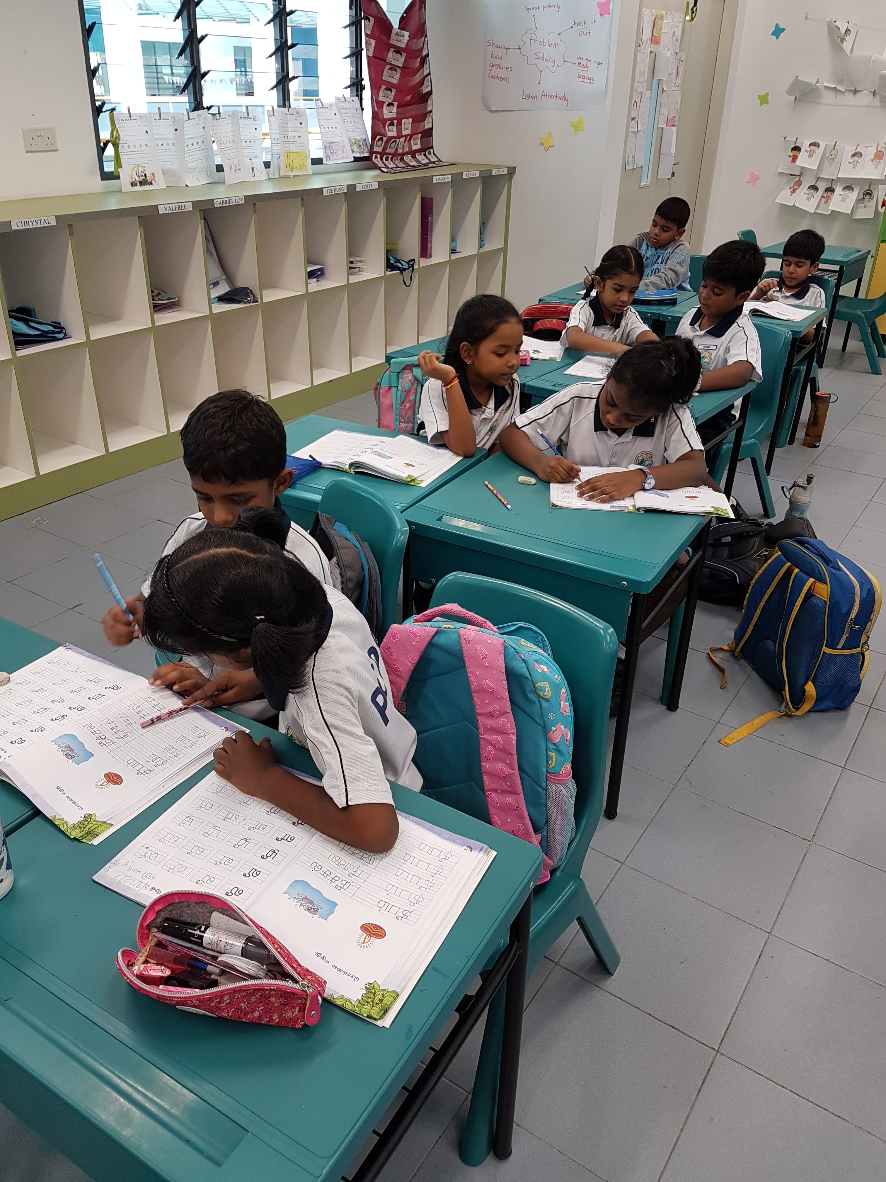 Children in school uniforms sit at teal desks, writing in workbooks; backpacks and cubbies line the room.