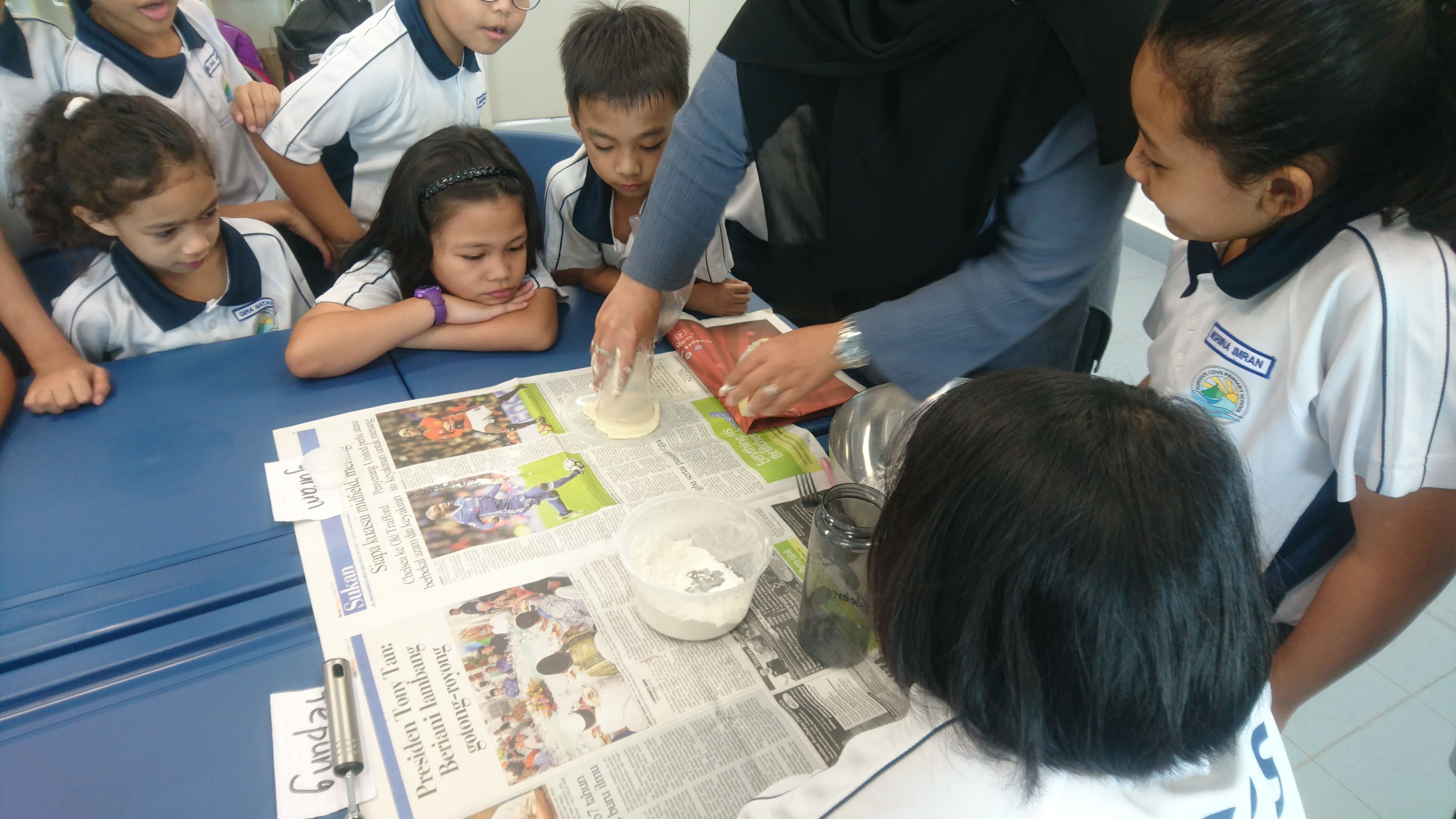 Children in school uniforms watch an adult knead dough on a newspaper-covered blue table with a bowl of flour.