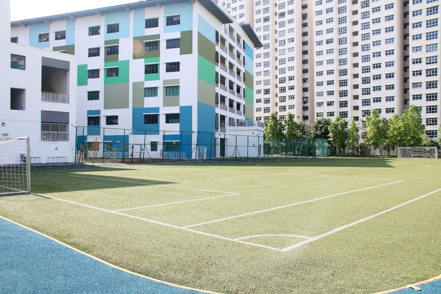An outdoor soccer field with artificial turf and goalposts, surrounded by colorful multi-story buildings and trees.