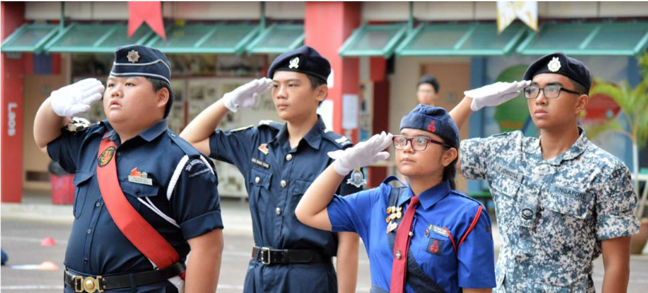Four young people in different uniforms and caps saluting outdoors near a red building.
