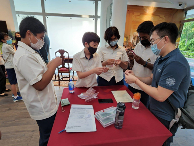 Students in white uniforms and face masks gather around a red table during an indoor hands-on activity.