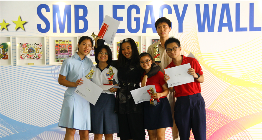Six students holding trophies and certificates pose in front of a colorful SMB Legacy Wall sign.