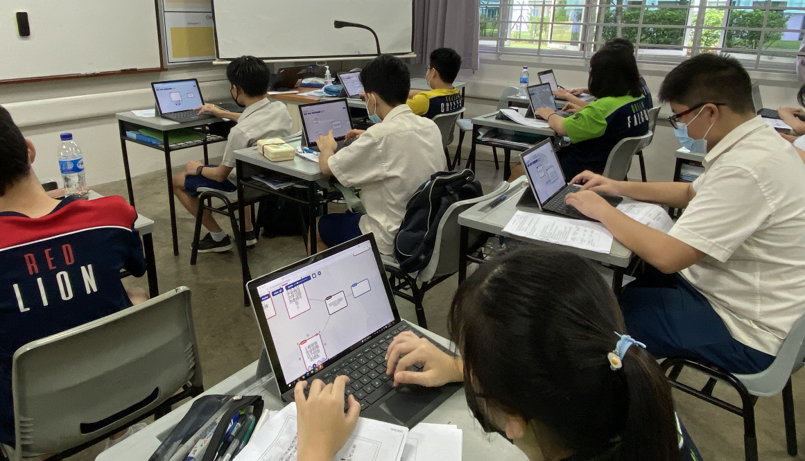 Students wearing masks sit at desks in a classroom, each working on a laptop with diagrams on screen.