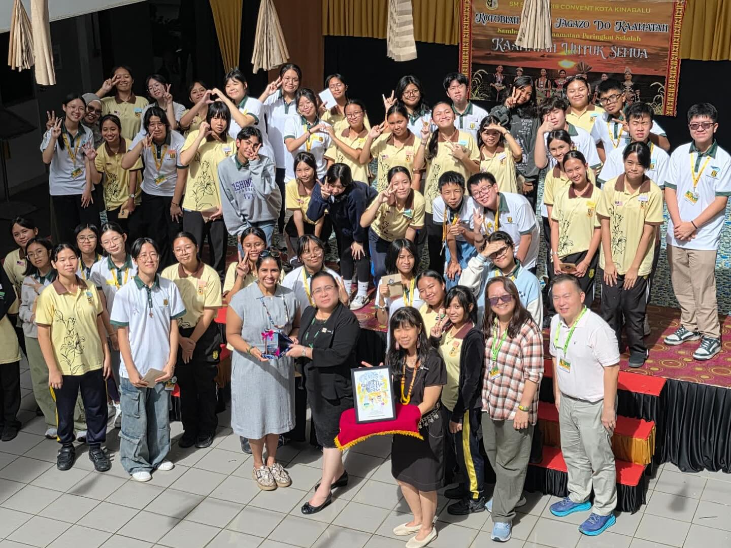 Large group of students and adults in yellow uniforms posing with peace signs at a school Kaamatan event in Kota Kinabalu.