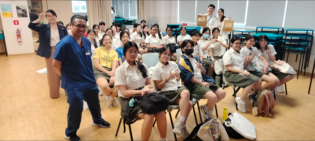 A man in blue scrubs poses with a large group of students in white uniforms seated in a classroom.