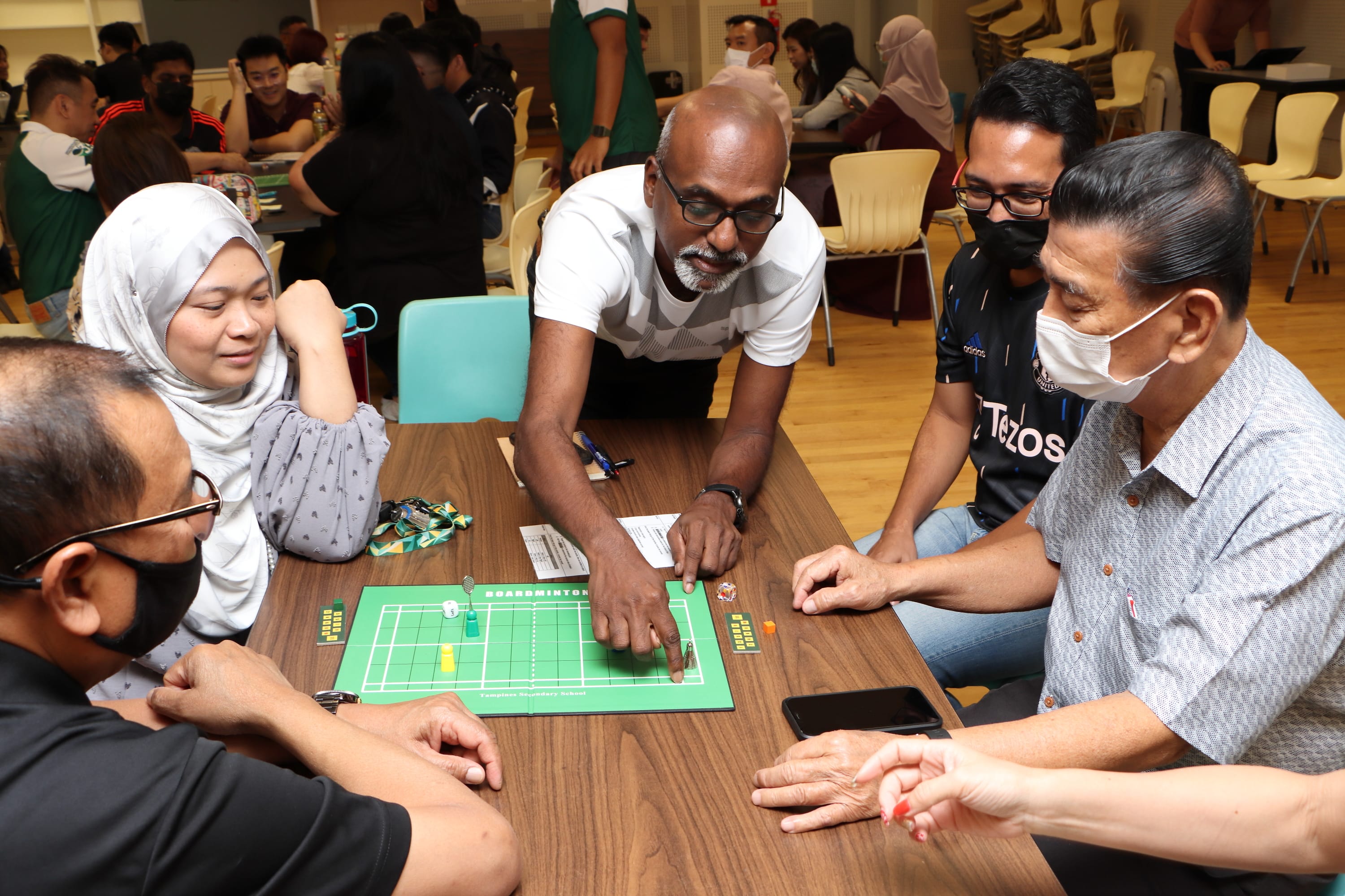 Group gathered at a table playing a badminton-themed board game, with one man pointing at the board.
