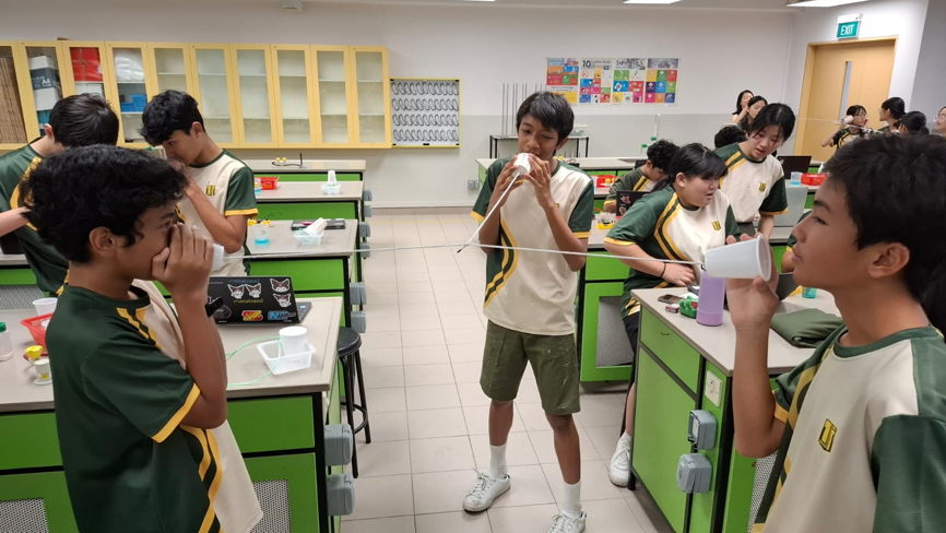 Students in uniforms use paper cup and string telephones in a school science laboratory.