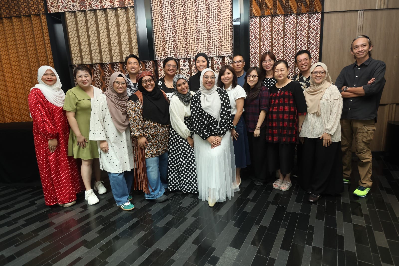 A large group of about 17 people posing together indoors in front of patterned batik fabric wall hangings.