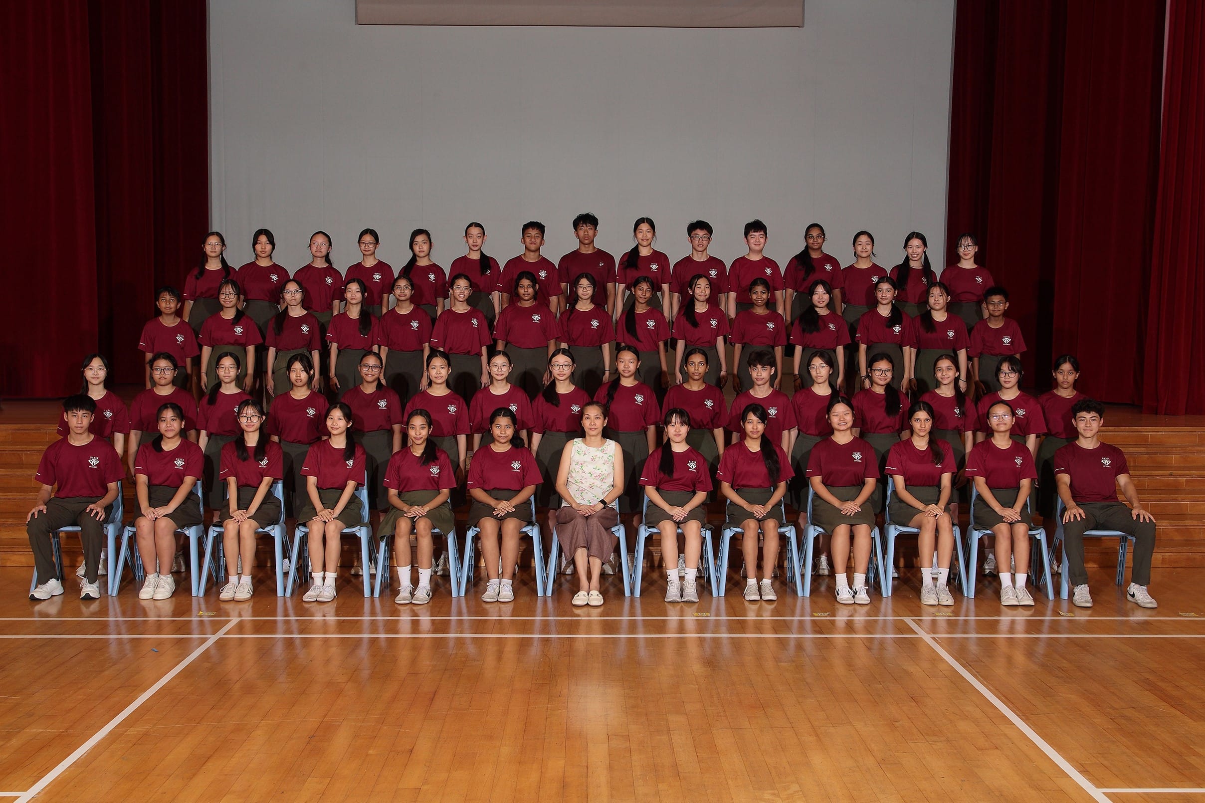 Large group of students in matching dark red uniforms posed in rows on a wooden school hall stage with one adult.