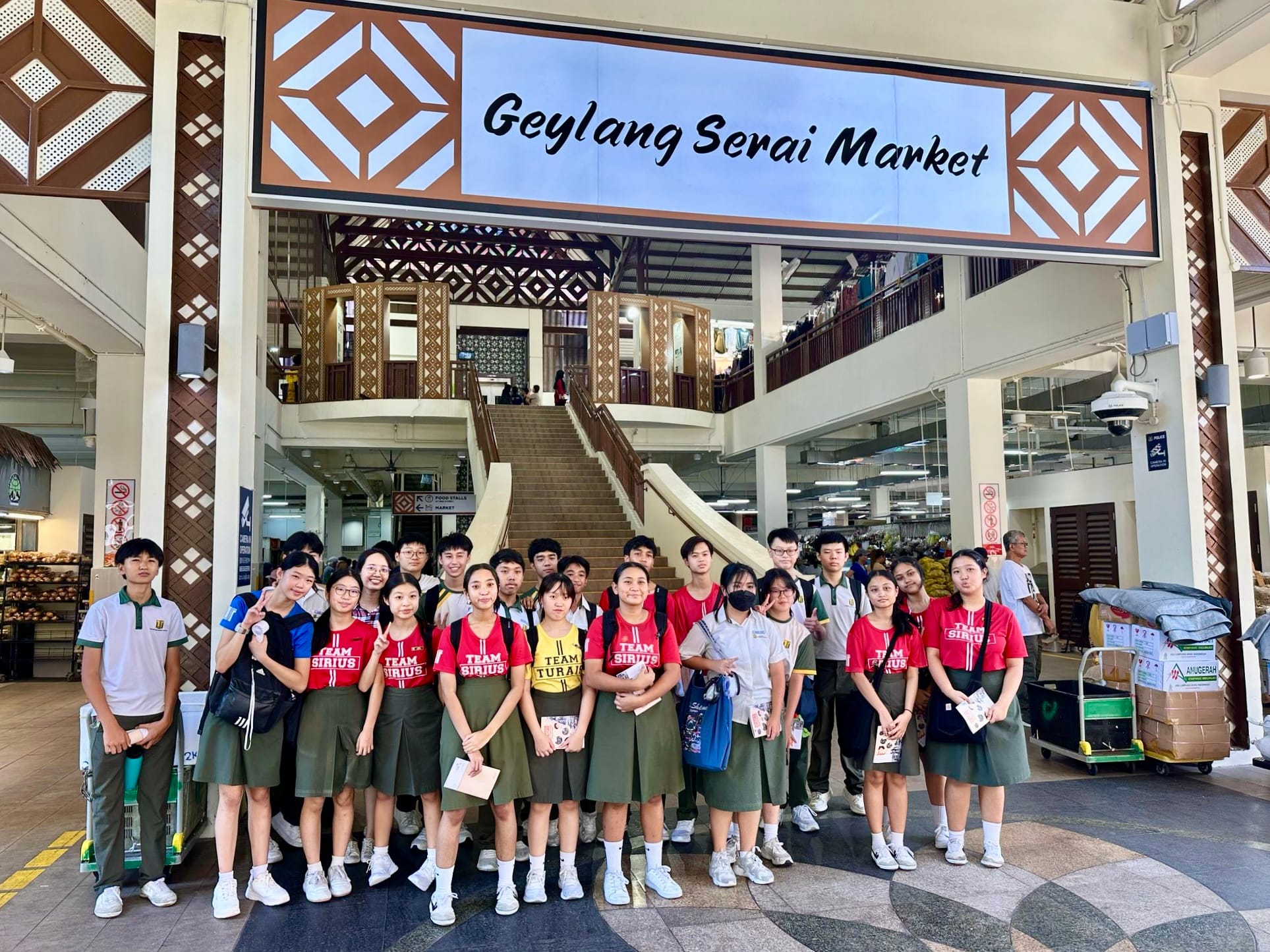A group of students in uniforms and red team shirts pose together at the entrance of Geylang Serai Market.
