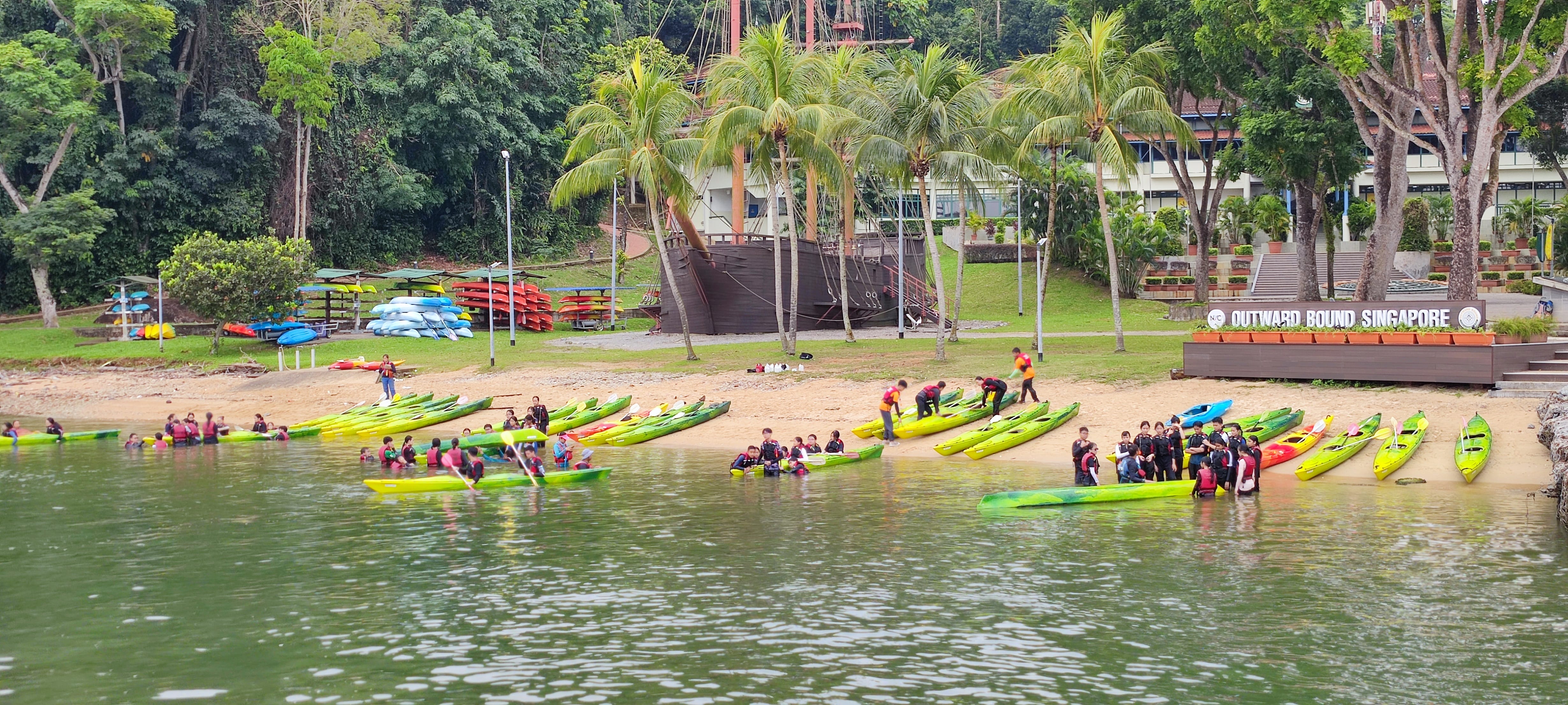 Groups in life jackets gather by green kayaks on a beach, with palm trees and buildings behind.