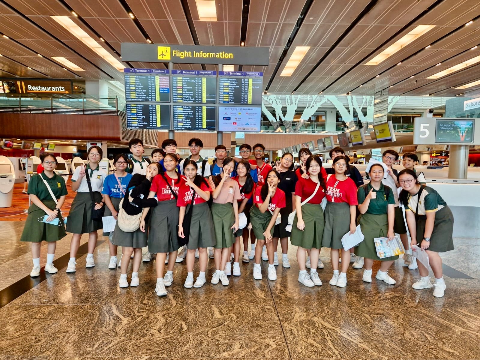 A large group of students in school uniforms pose under a Flight Information board at an airport terminal.