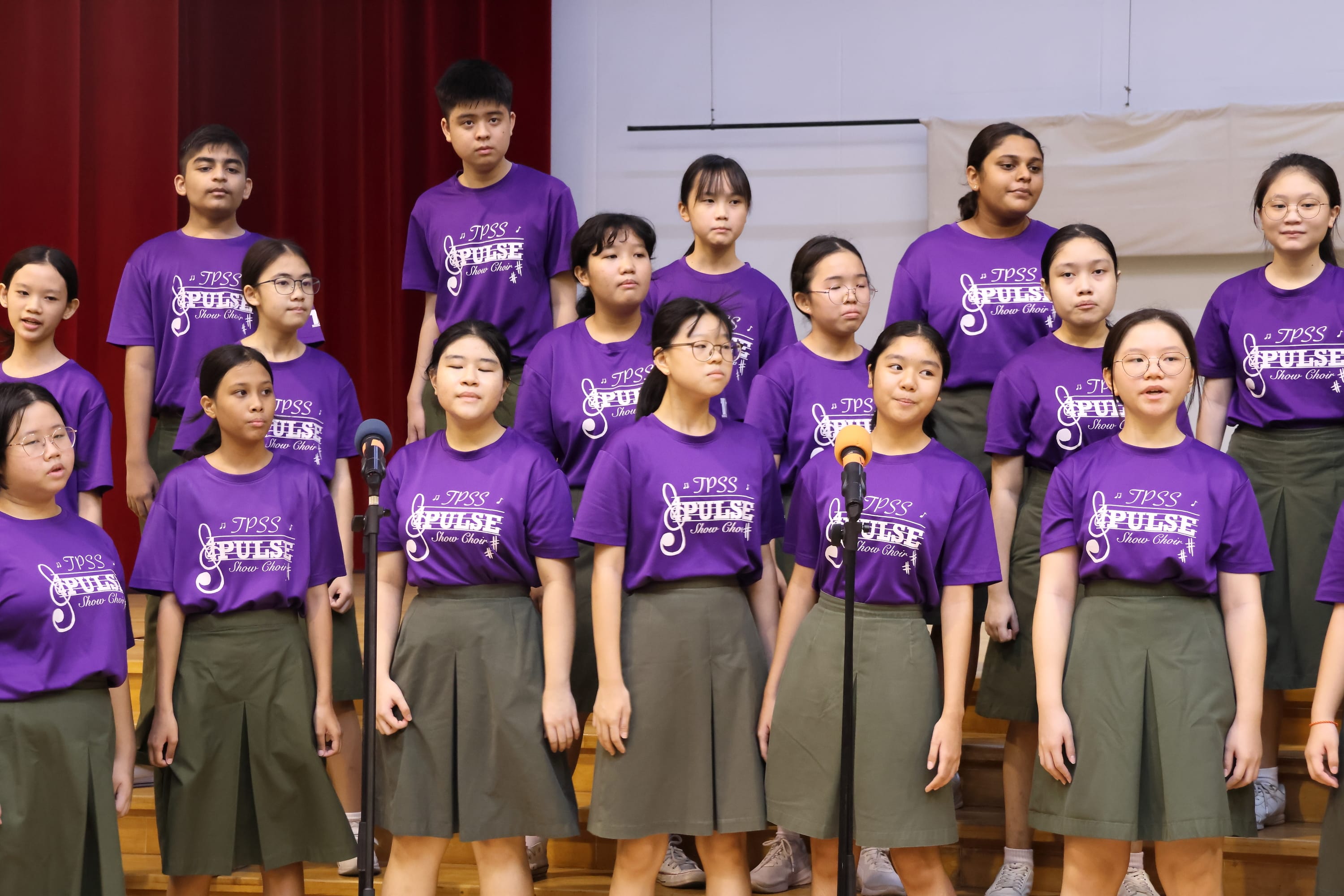 Choir of students in purple shirts and olive skirts on stage risers, with two microphones in front.