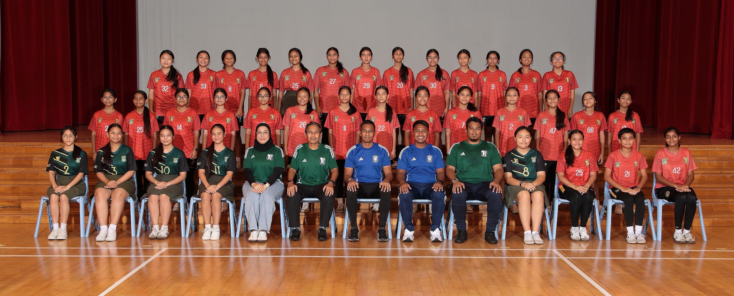 A large group of young female soccer players in red jerseys with coaches in green and blue, posing in three rows indoors.