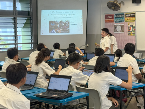 Students in white uniforms sit at desks with laptops facing a presenter and a projected slide at the front.