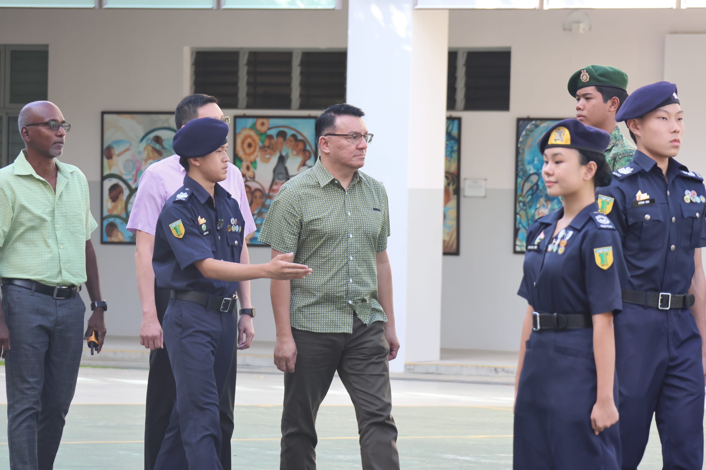 Uniformed cadets march past as a cadet gestures while walking with three men in a courtyard.