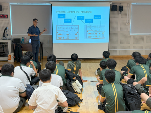 A man presents a projector controller patch panel slide to students in green uniforms seated on a floor.