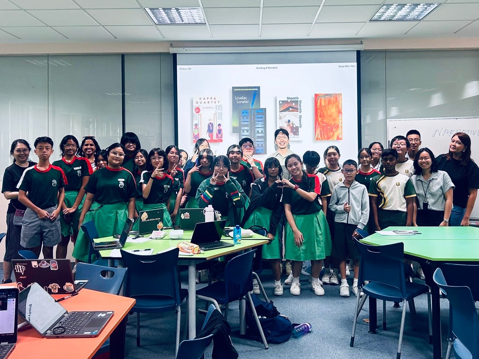 A large group of students in green uniforms pose together in a classroom with a Fiction 101 presentation on a projector screen.