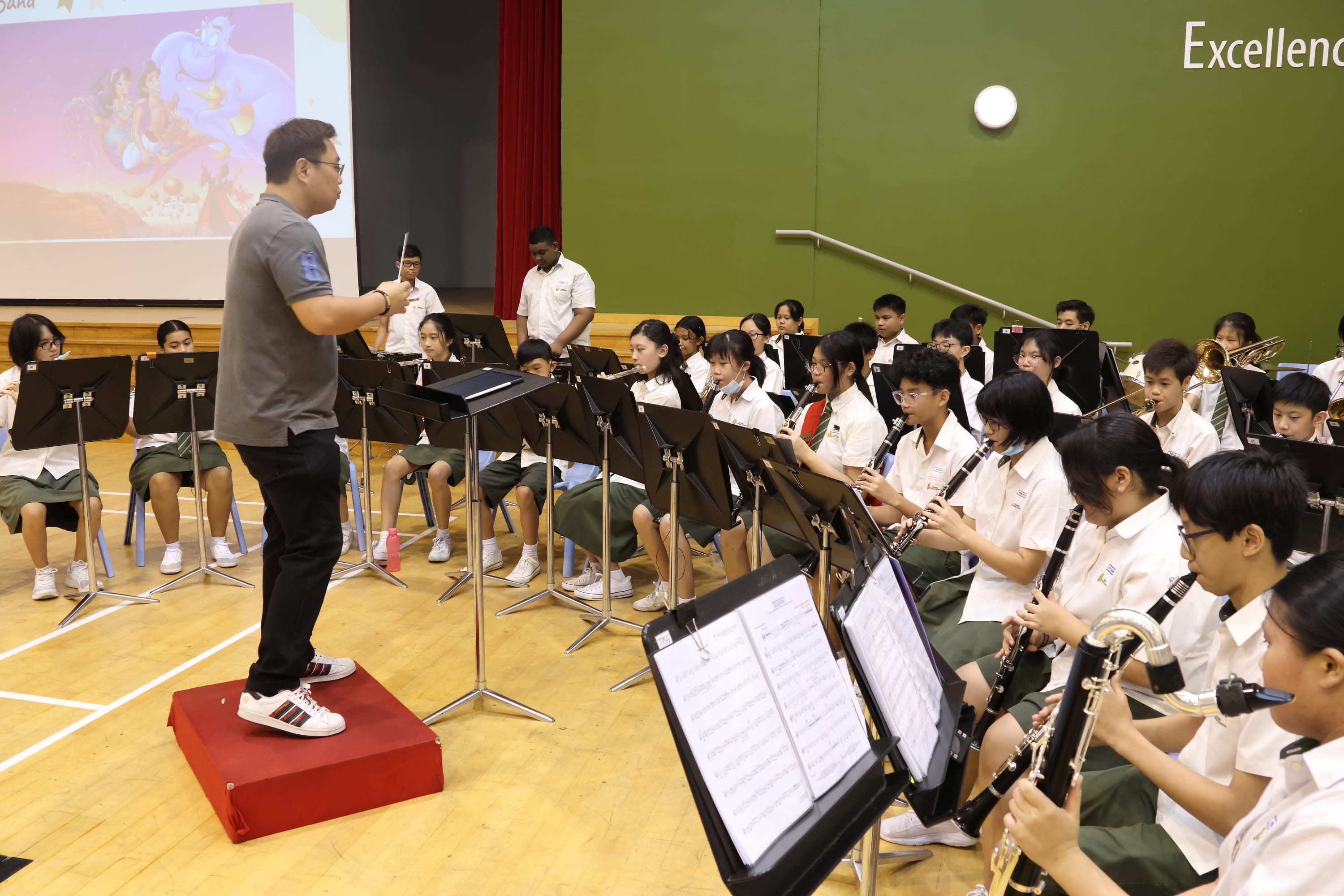 Conductor stands on a red podium leading a seated student wind band with music stands in a hall.