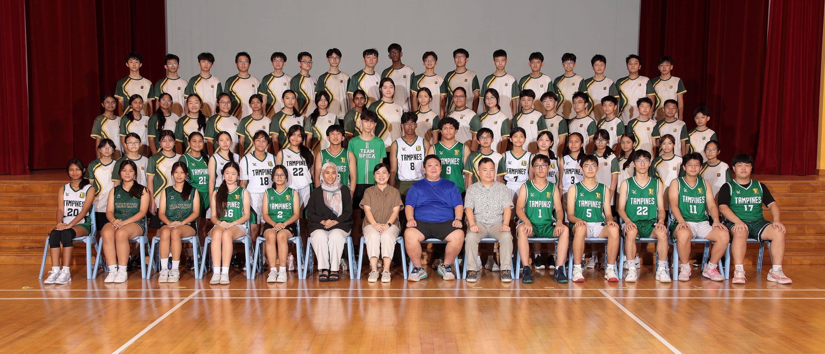Large group of Tampines school basketball team members and coaches posing in green and white uniforms indoors.