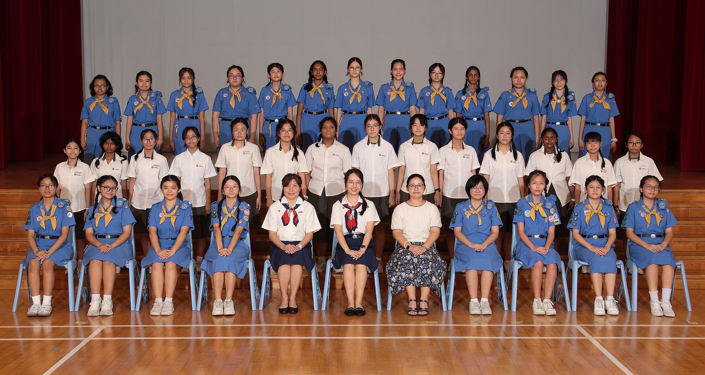 Large group of girls in blue scout uniforms with yellow neckerchiefs posing in rows with adult leaders seated in front.