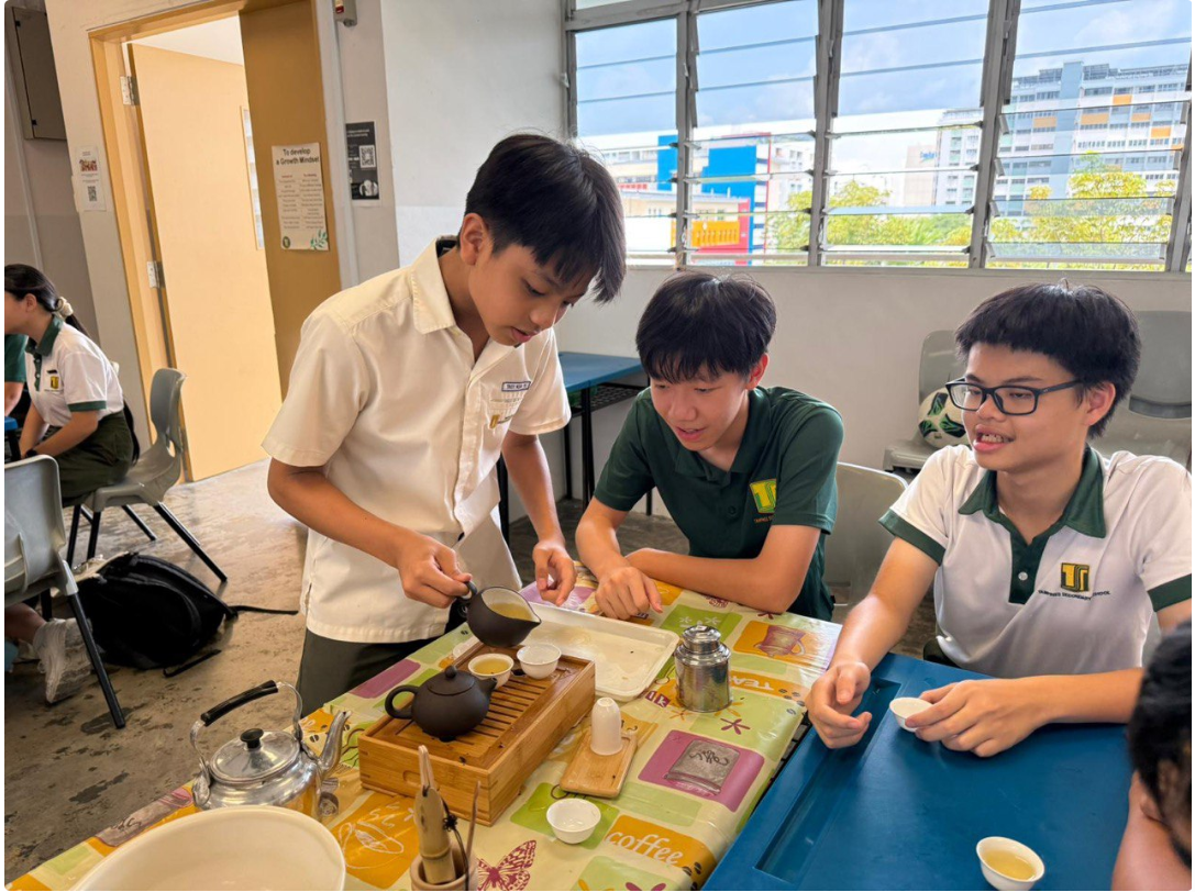 Three school students in uniforms practice a traditional tea ceremony with a clay teapot and wooden tray on a table.