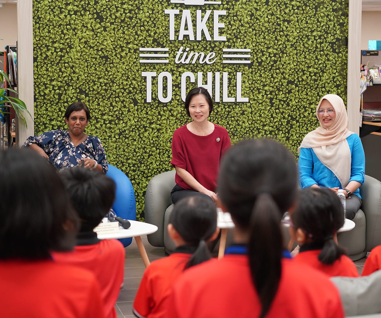 Three women sit before a green wall reading "Take Time To Chill," facing a group of children in red uniforms.